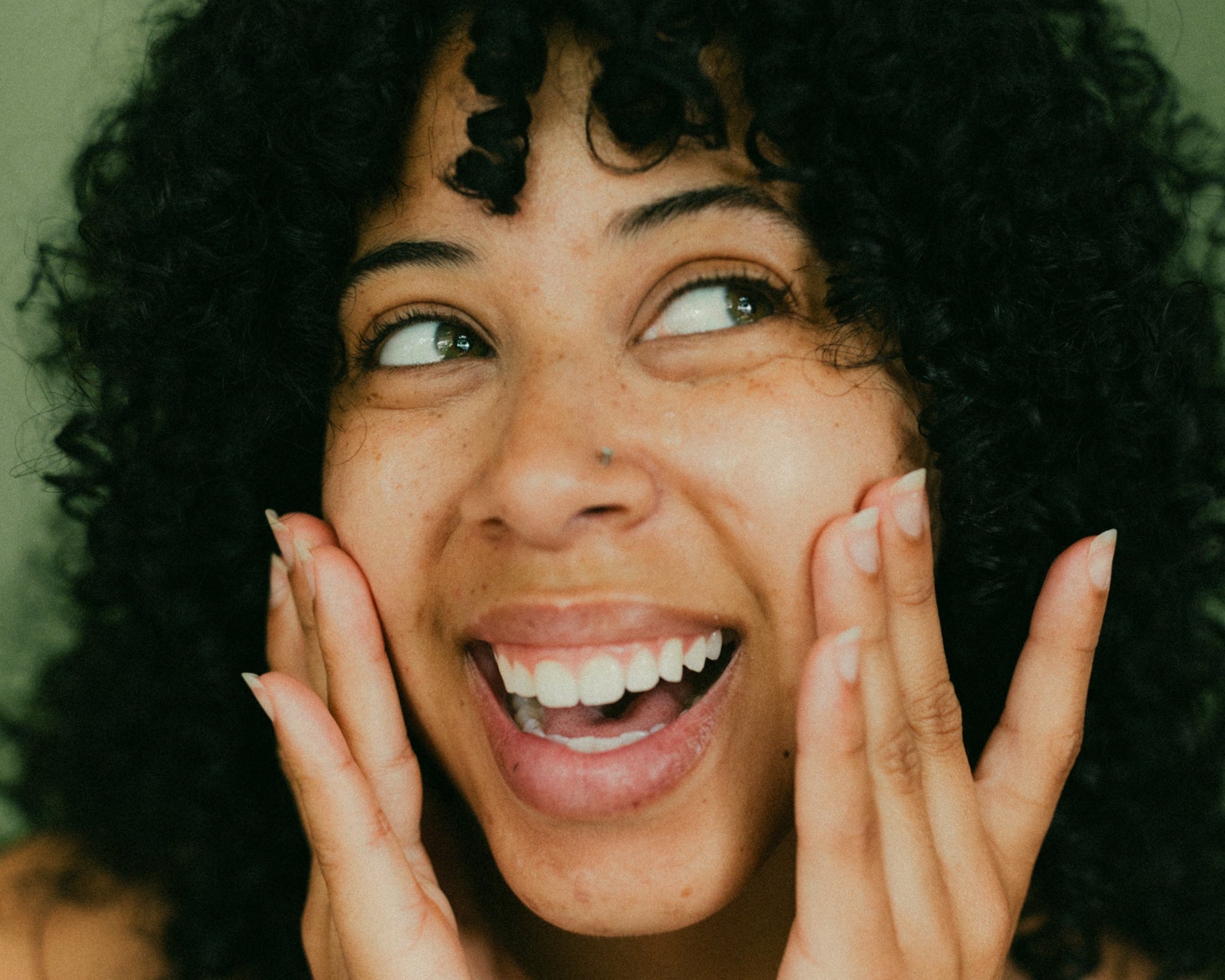 A young woman with curly black hair smiling brightly and touching her face with both hands against a green background.