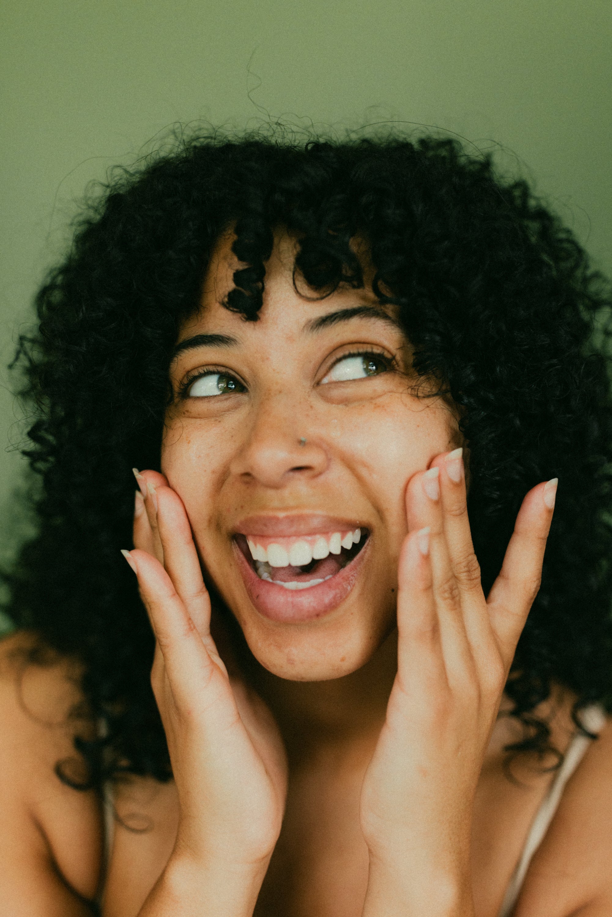 A young woman with curly black hair smiling brightly and touching her face with both hands against a green background.