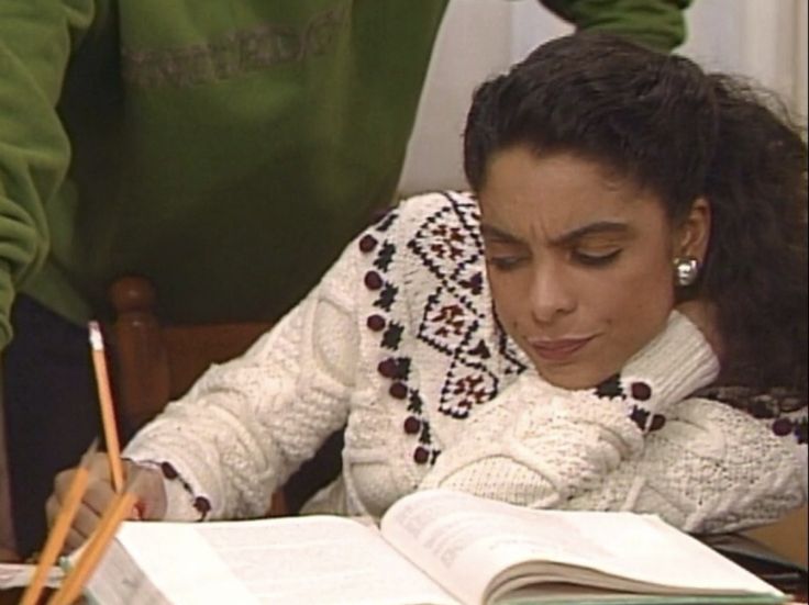 A young woman in a patterned white cable-knit sweater leans her head on her hand, looking focused and stressed while studying a large open textbook at a desk.