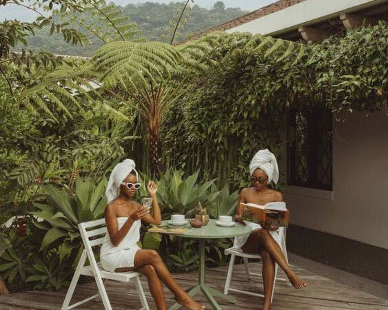 Two women wrapped in white bath towels relax at an outdoor table on a wooden patio surrounded by tropical plants.