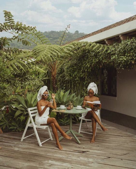 Two women wrapped in white bath towels relax at an outdoor table on a wooden patio surrounded by tropical plants.