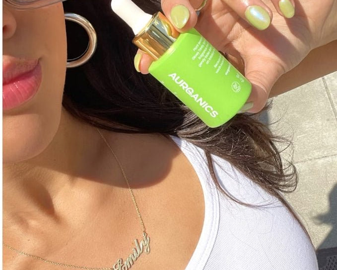 A woman wearing a white tank top holds up a bright green skincare bottle by Aurganics that matches her light green nail polish.