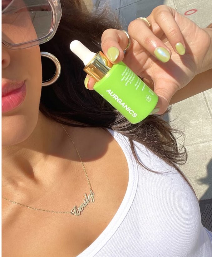 A woman wearing a white tank top holds up a bright green skincare bottle by Aurganics that matches her light green nail polish.