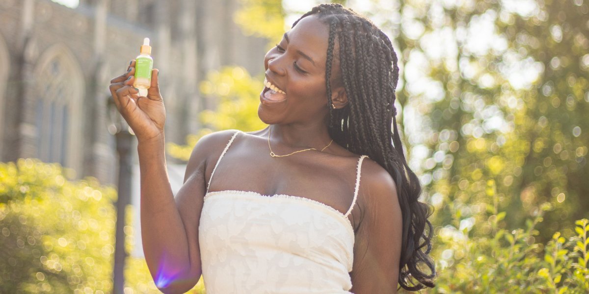 A smiling young Black woman holding up a small skincare dropper bottle while standing outside in sunny weather.