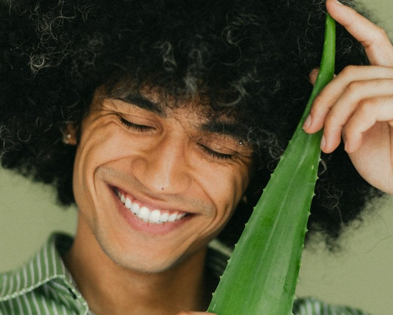 A young man with a curly afro smiles warmly while holding up a large aloe vera leaf next to his face against a green background.