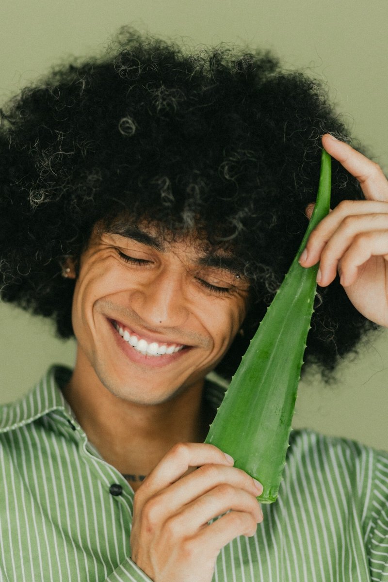 A young man with a curly afro smiles warmly while holding up a large aloe vera leaf next to his face against a green background.