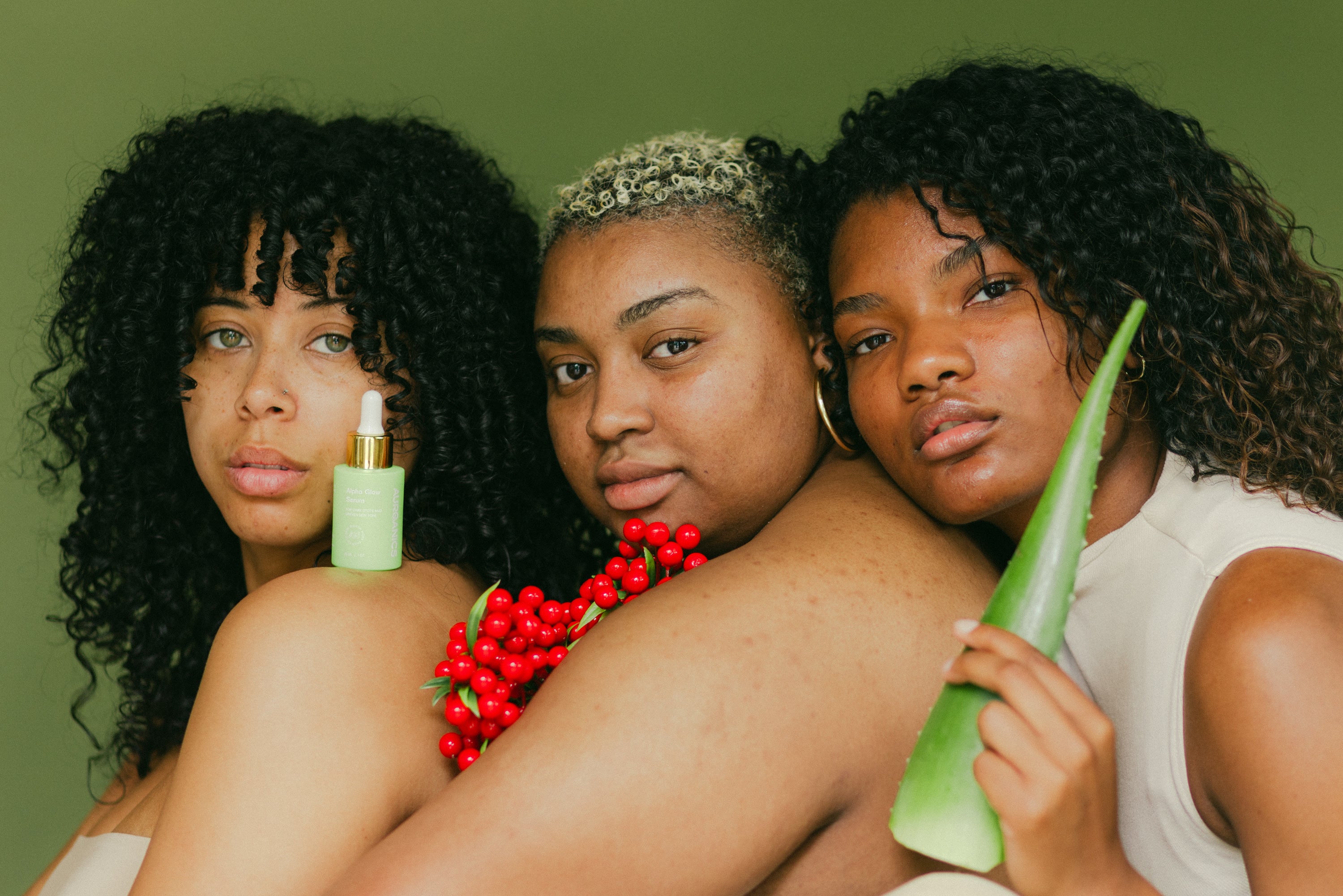 Three women with melanated skin holding aloe vera and alpha arbutin ingredients for hyperpigmentation treatment in the Alpha glow serum