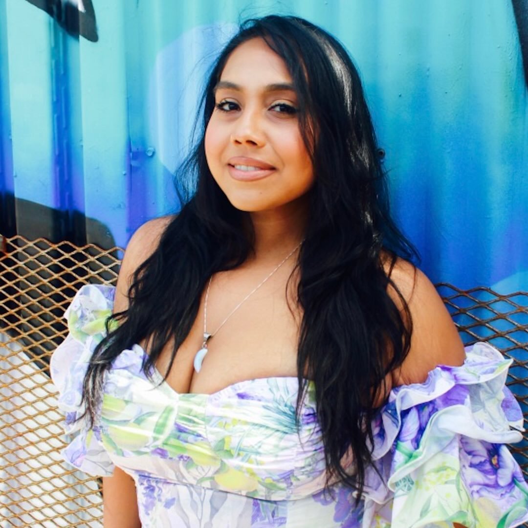 Woman wearing a colorful floral dress sitting against a blue wall.