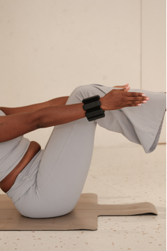 A person in light gray athletic wear performing a yoga or exercise pose on a beige mat, wearing a black wrist strap device on one wrist.